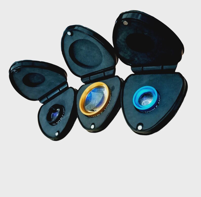 Puede incluir: Tres estuches negros en forma de l&aacute;grima con tapas abatibles, cada uno con una lente circular. La lente izquierda es negra, la central tiene un borde dorado y la derecha un borde azul brillante. Cada estuche incluye un inserto de espuma.