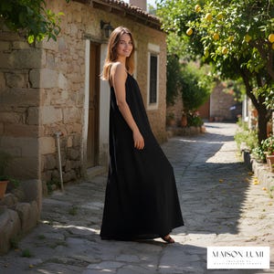 May include: A black maxi dress with thin straps. The dress is worn by a person on a cobbled street. The background features a stone building, trees, and the text "MAISON LUMII".