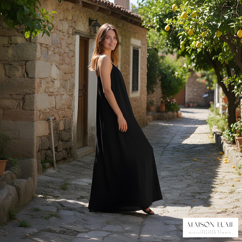 May include: A black maxi dress with thin straps. The dress is worn by a person on a cobbled street. The background features a stone building, trees, and the text "MAISON LUMII".