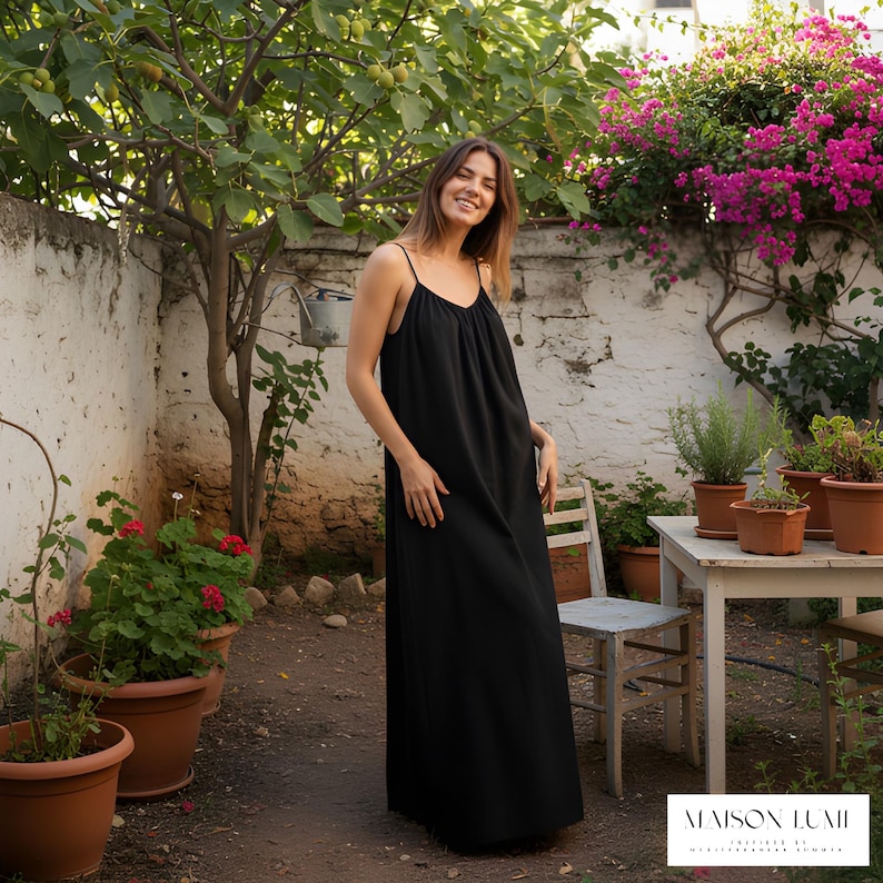 May include: A black maxi dress with thin straps. The dress is worn in a garden with potted plants and a white wall. The dress has a simple, flowing design. The brand name "MAISON LUMI" is visible in the bottom right corner.