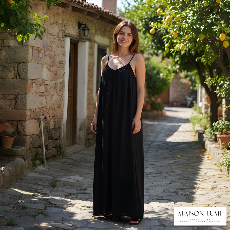 May include: A black maxi dress with thin straps is worn by a woman on a cobbled street. The dress is long and flowing. The background includes stone buildings, lemon trees, and the text "MAISON LUMII".