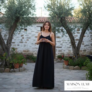May include: A woman in a black maxi dress with thin straps, holding a mug. The dress is floor-length. The background includes a stone building, olive trees, and potted plants. The text "MAISON LUMI" is visible at the bottom.