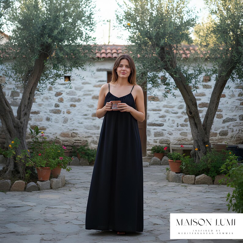 May include: A woman in a black maxi dress with thin straps, holding a mug. The dress is floor-length. The background includes a stone building, olive trees, and potted plants. The text "MAISON LUMI" is visible at the bottom.