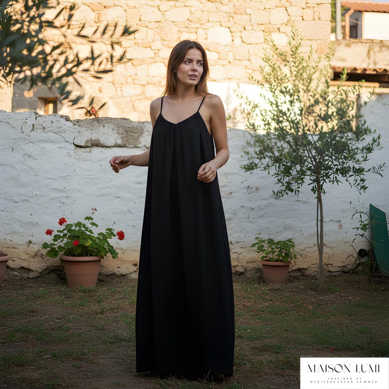 May include: A black maxi dress with thin straps. The dress has a loose, flowing design and a V-neck. The model is outdoors, standing in front of a white wall and green grass. The dress is from Maison Lumii.