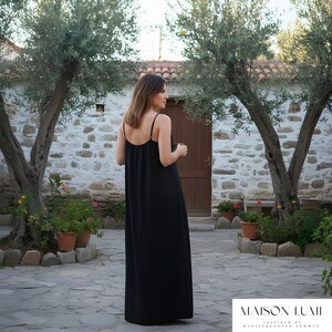 May include: A black maxi dress with thin straps. The dress is worn in a courtyard with olive trees and stone walls. The brand name "MAISON LUMI" is displayed at the bottom of the image. The dress is floor-length.