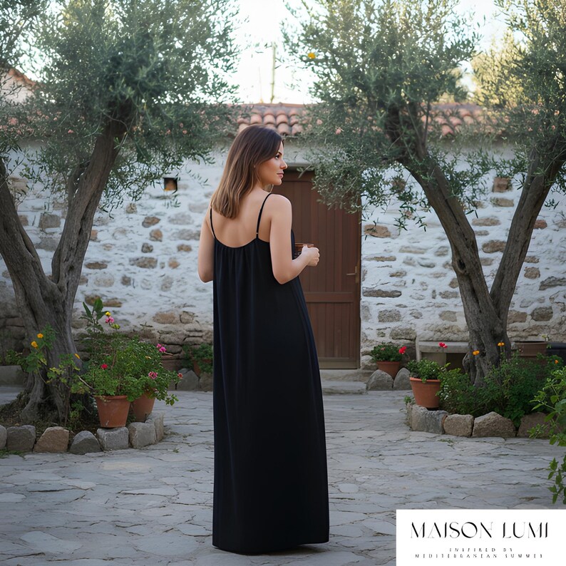 May include: A black maxi dress with thin straps. The dress is worn in a courtyard with olive trees and stone walls. The brand name "MAISON LUMI" is displayed at the bottom of the image. The dress is floor-length.
