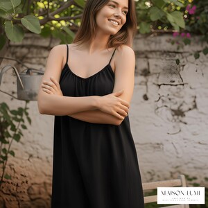 May include: A black spaghetti strap dress with a V-neck. The dress is worn by a woman. The background features a white stone wall, greenery, and a watering can. The dress has a flowing silhouette.