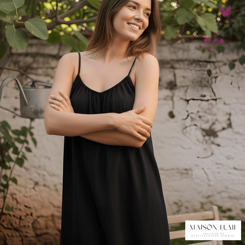 May include: A black spaghetti strap dress with a V-neck. The dress is worn by a woman. The background features a white stone wall, greenery, and a watering can. The dress has a flowing silhouette.