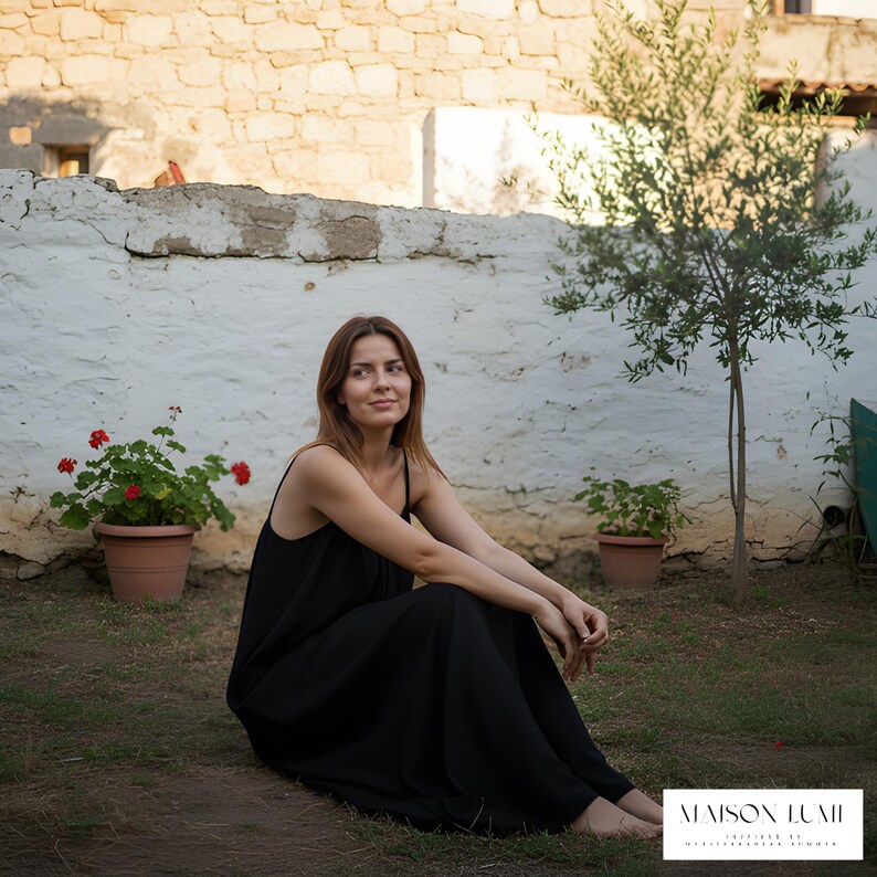 May include: A woman in a black maxi dress sits on grass, with a white stone wall behind her. Red flowers in a terracotta pot are on the left. A small tree and the text "MAISON LUMI" are also visible.