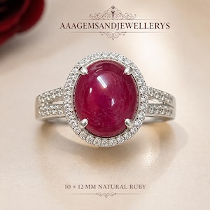 Könnte beinhalten: Ein silberner Ring mit einem großen ovalen, im Cabochon-Schliff gehaltenen natürlichen Rubin von 10 x 12 mm, umgeben von einem Kranz aus kleinen weißen Steinen. Die Ringschiene ist geteilt und mit kleinen weißen Steinen besetzt. Text: AAAGEMSANDJEWELLERYS.