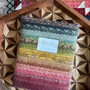 Può includere: Un fascio di strisce di tessuto in vari colori, tra cui verde, giallo, rosa e bordeaux, con motivi floreali e geometrici. Il tessuto è confezionato in plastica trasparente e ha un'etichetta che recita "STITCHED by Starlight". Il fascio è su un vassoio di legno.