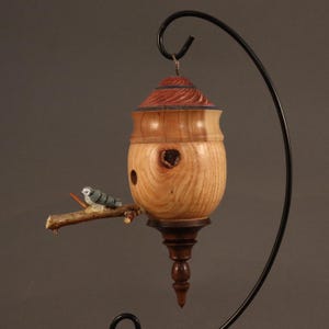 May include: A handcrafted wooden birdhouse with a decorative bird perched on a branch. The birdhouse features a natural wood grain finish, a red and brown top, and a dark brown base. The bird is painted in shades of gray and blue. The birdhouse is suspended from a black metal stand.