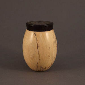 May include: A handcrafted wooden jar with a black lid. The jar has a light-colored body with dark streaks and a rounded, egg-like shape. The lid is a solid, dark color, contrasting with the lighter wood.