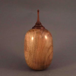 May include: A handcrafted wooden urn with a dark brown lid and a light brown body. The urn has a smooth, polished finish and a tall, pointed finial on top. The wood grain is visible, adding to its natural aesthetic.