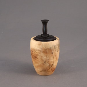 May include: A small wooden box with a natural wood grain finish and a black wooden lid with a knob. The box is oval shaped and has a smooth surface.