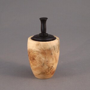 May include: A small, handcrafted wooden jar with a lid. The jar's body is light-colored wood with a natural grain pattern. The lid and handle are dark brown. The jar has a rounded shape and stands on a gray background.