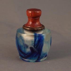 May include: A clear glass bottle with a blue and white swirl design and a wooden top. The bottle has a round shape and a small opening at the top.