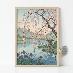 以下が含まれることがあります： ピンクの花を咲かせた桜の木が穏やかな青い池の上に枝を広げる風景を描いた額装アートプリント。草の生い茂る岸辺には石灯籠と紫色のアイリスが配置されている。絵の中には日本語の書体による文字が描かれている。