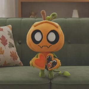Puede incluir: Un peluche con forma de personaje con cabeza de calabaza, con grandes ojos blancos y boca cosida. Tiene brazos y piernas verdes y sostiene dos objetos en forma de corazón. El peluche es naranja y verde, y está sobre un sofá verde.