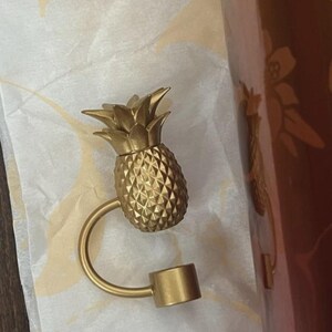 May include: A gold-colored metal bottle stopper featuring a textured pineapple-shaped top. The pineapple is attached to a curved metal arm that connects to a cylindrical base designed to fit into the opening of a bottle for sealing purposes.