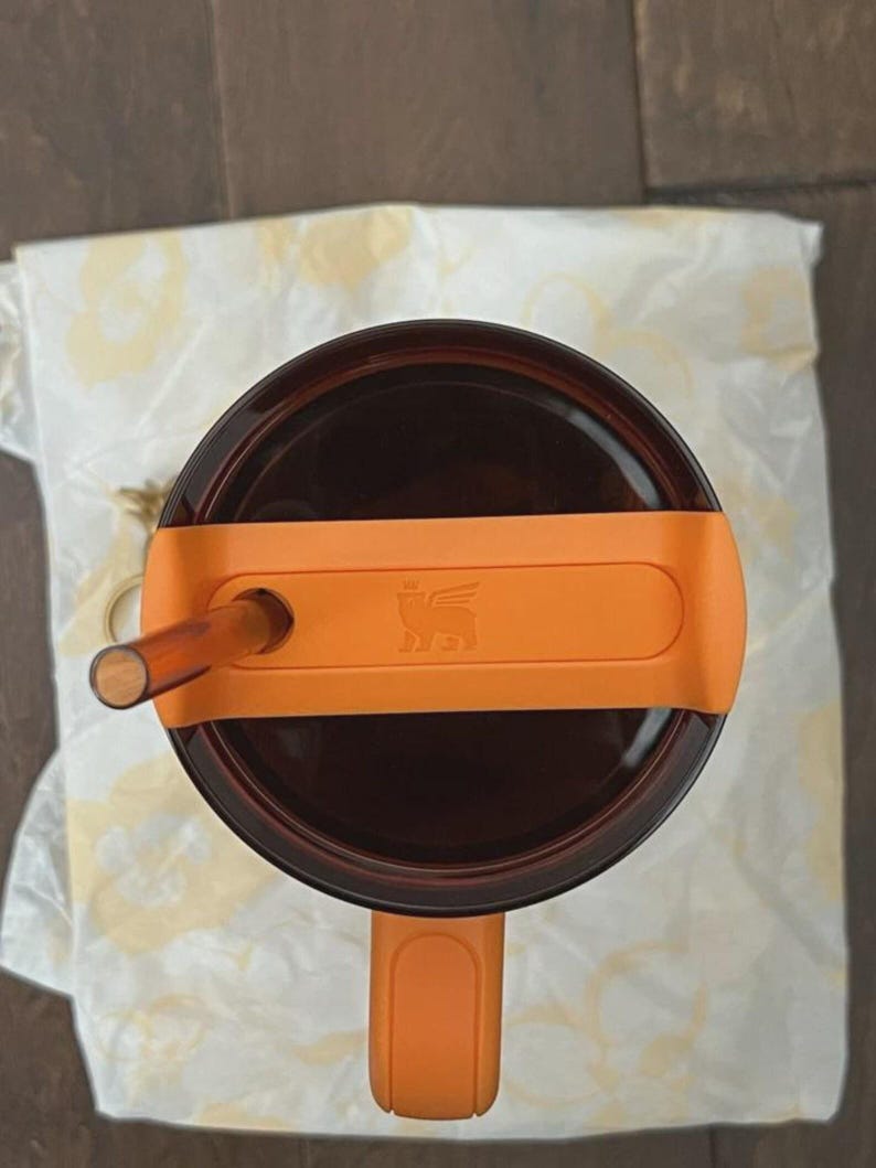May include: A top-down view of a dark brown translucent tumbler featuring a bright orange lid and a matching orange handle. A dark brown straw protrudes from the lid, which displays a small embossed logo in the center of the orange plastic surface.
