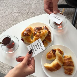 Op de afbeelding: Een witte tafel met borden met goudbruine gebakjes en heldere glazen thee. Vraagkaarten worden boven de tafel gehouden. Eén kaart luidt: "Welke angsten heb je over culturele verschillen in onze..." en een andere "Question Card."