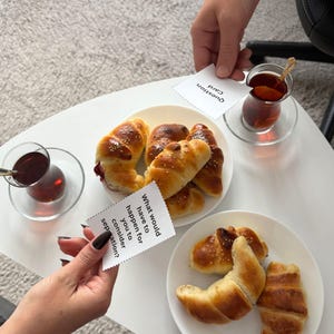 May include: A white table setting with plates of baked goods, including croissants and rolls. Two glasses of tea are present, along with question cards. One card reads "What would have to happen for you to consider separation?" and another reads "Question Card."