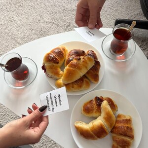 Puede incluir: Dos platos blancos con productos horneados, incluyendo cruasanes y panecillos, están sobre una mesa blanca. Dos vasos de té están sobre platillos. Pequeñas tarjetas con texto son sostenidas por manos. El texto en una tarjeta dice: "Welche Träume musstest du loslassen?"