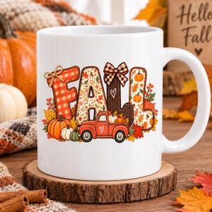 Puede incluir: Una taza de cerámica blanca con la palabra FALL en letras grandes. La F es de cuadros rojos, la A y la L están rellenas de hojas de otoño, y la segunda L es marrón con un lazo de cuadros. Una camioneta roja con calabazas bajo las letras.