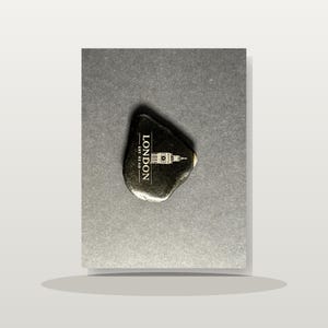 May include: A smooth, dark, irregularly shaped stone featuring a white printed graphic of the Big Ben clock tower and the text LONDON EST. 43 AD. The stone has a polished, reflective surface and is centered on a dark gray rectangular card.