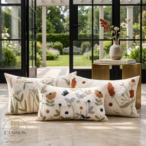 May include: Three rectangular throw pillows with an off-white base featuring embroidered botanical designs of various flowers in shades of white, orange, and blue, including a small blue butterfly detail on the center pillow and a logo in the corner.