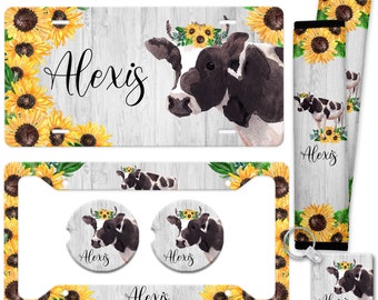 May include: A set of car accessories featuring a black and white cow with a sunflower crown. The set includes a license plate frame, two coasters, a key chain, and two seat belt covers. The items are all decorated with sunflowers and the name "Alexis".