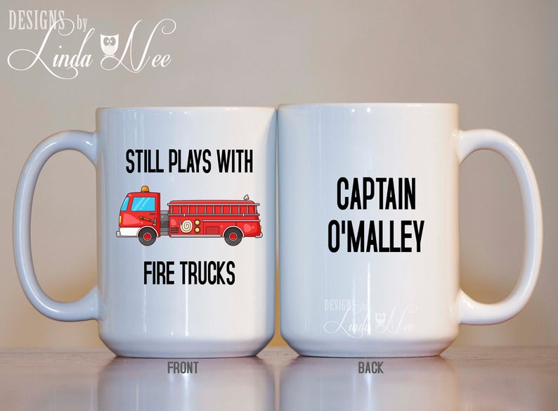 Peut inclure: Une tasse &agrave; caf&eacute; en c&eacute;ramique blanche avec une illustration de camion de pompiers rouge et le texte "Still Plays With Fire Trucks" sur le devant. Le dos de la tasse porte le texte "Captain O'Malley".