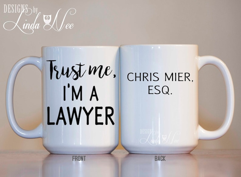 K&ouml;nnte beinhalten: Wei&szlig;e Keramik-Kaffeetasse mit schwarzem Text, der auf der Vorderseite "Vertrau mir, ich bin Anwalt" und auf der R&uuml;ckseite "Chris Mier, Esq." lautet.