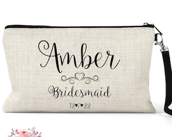May include: A beige linen makeup bag with a black zipper and a black strap. The bag has the name "Amber" written in cursive with a heart design and the word "Bridesmaid" written below. The date "12 7 22" is written below the word "Bridesmaid".