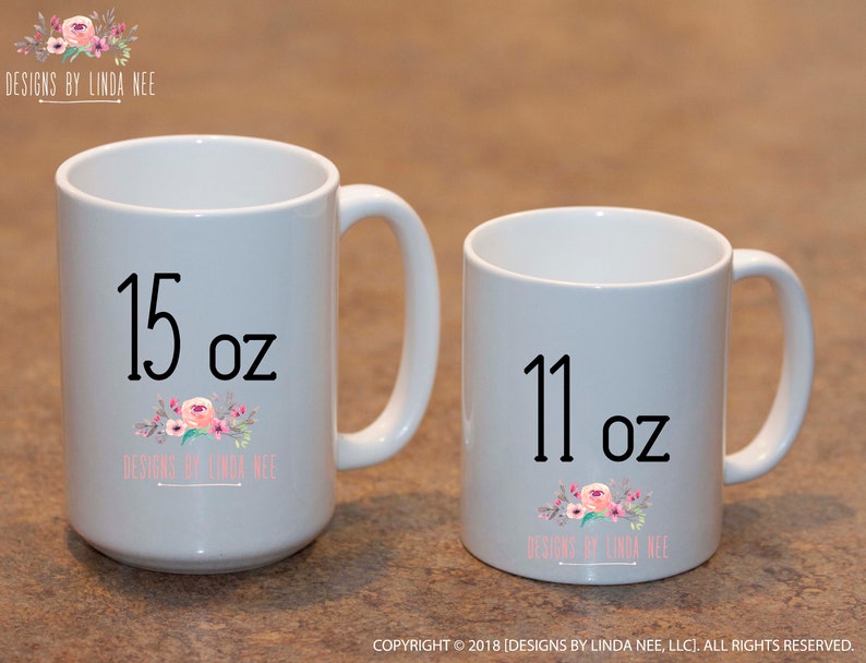 May include: Two white ceramic mugs. The larger mug is labeled "15 oz" and the smaller mug is labeled "11 oz". Both mugs feature a floral design and the text "DESIGNS BY LINDA NEE".