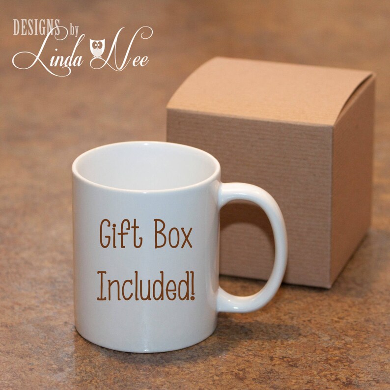 K&ouml;nnte beinhalten: Eine wei&szlig;e Keramiktasse mit braunem Henkel. Auf der Tasse steht in braunen Buchstaben "Gift Box Included!" gedruckt. Die Tasse steht auf einer braunen Oberfl&auml;che neben einer braunen Pappgeschenkbox.