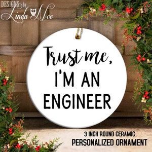 Könnte beinhalten: Runder weißer Keramik-Anhänger mit schwarzem Text, der "Trust me, I'M AN ENGINEER" lautet. Der Anhänger hat einen Durchmesser von 7,6 cm.