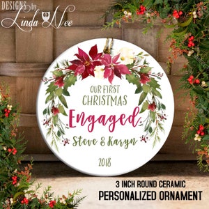 Puede incluir: Un adorno redondo de cerámica blanca con un diseño de corona de flores de acuarela en tonos verdes, rojos y blancos. El adorno presenta el texto "Our First Christmas Engaged Steve & Karyn 2018". El adorno tiene un diámetro de 7,6 cm.