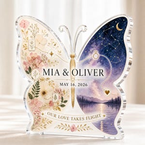 Pode incluir: Uma placa de acrílico em forma de borboleta com design dividido. O lado esquerdo exibe flores cor-de-rosa prensadas e folhagem. O lado direito mostra um céu noturno com estrelas, uma lua crescente e um lago. Texto: MIA & OLIVER, MAY 16, 2026, OUR LOVE TAKES FLIGHT.