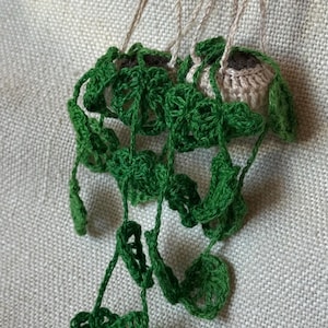 Puede incluir: Un par de adornos de plantas colgantes de ganchillo con hojas de color verde oscuro que cuelgan de pequeñas macetas beige. Cada maceta está suspendida por finos cordones de hilo beige unidos a una anilla de metal plateado como decoración.