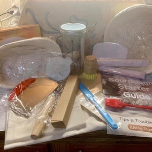 May include: A collection of sourdough bread-making tools, including a banneton basket, a dough scraper, a bench scraper, a jar, a brush, a scale, and a guide. The items are arranged on a wooden surface, with a white cloth and a pillow in the background.