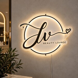 以下が含まれることがあります： バックライト付きの円形壁掛け看板。黒い筆記体の文字「lv」とサンセリフ体の「BEAUTY LOUNGE」というテキストが記されています。筆記体の右側には小さな黒いハートのアイコンがあります。看板の表面は白です。