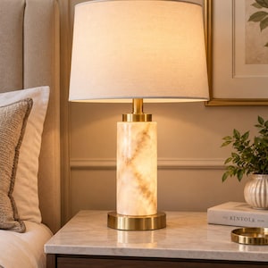 May include: A table lamp featuring a cylindrical base made of white and beige marbled stone, accented with a brushed gold metal base and neck. The lamp is topped with a large, tapered off-white fabric drum shade and a small gold finial.