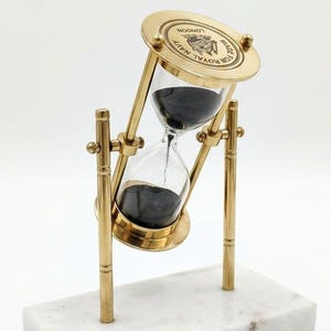 May include: A brass hourglass with black sand, mounted on a rotating frame supported by two vertical brass pillars. The top brass plate is engraved with the text "MADE FOR ROYAL NAVY LONDON". The entire structure rests on a rectangular white marble base.