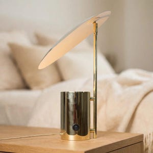 May include: A gold-colored metal desk lamp featuring a cylindrical base with a black power switch and a thin vertical stem supporting a large, flat, circular shade angled downward to provide illumination from its underside surface.