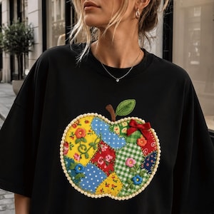 Op de afbeelding: Een zwart T-shirt met korte mouwen en een grote appelafbeelding in het midden. De appel is samengesteld uit diverse kleurrijke stoffen lapjes met bloemen- en stippenpatronen, omlijnd door een rand van kleine witte parels en een rode strik.