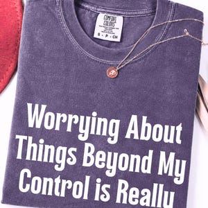 May include: A purple short-sleeve t-shirt with a crew neckline and a white graphic print that reads, "Worrying About Things Beyond My Control is Really". The shirt features a Comfort Colors brand label at the inner neck and a rose pendant necklace.