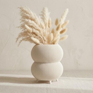 Peut inclure: Un vase en céramique couleur crème avec une texture côtelée à double bulbe et de petits pieds, rempli d'un grand arrangement d'herbes de la pampa beiges et duveteuses et de tiges de queue de lapin séchées s'étendant vers le haut.