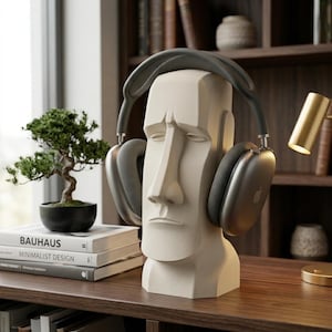 Puede incluir: Un soporte para auriculares con forma de cabeza Moái blanca con auriculares grises. El soporte está sobre una estantería de madera con un pequeño bonsái y libros. Los auriculares tienen el logotipo de Apple plateado en los auriculares.